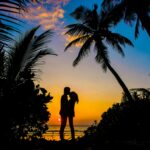 Silhouette of a couple sharing a romantic moment on a tropical beach at sunset with palm trees.